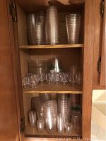 Shelves inside a wooden cabinet with multiple stacks of clear plastic cups including tall party cups, smaller cups, and shot glasses.