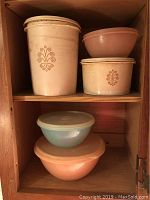 Five vintage Tupperware pieces stacked inside an open shelving unit. Two large cylindrical containers with lids and floral designs on top shelf, plus two nested mixing bowls below, one without lid.