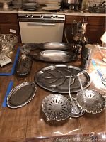 Photo shows multiple silver plate and stainless steel platters and a coffee urn arranged on a table. Visible are two elongated stainless steel platters, a large ornate silver divided platter, the coffee urn, and other smaller silver plate trays.