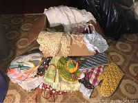 Box containing assorted vintage doilies, a white lacy table runner, and various other linens and pot holders stacked inside and around the box.