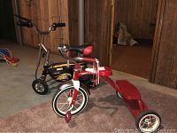 Two toddler bikes, one red and white Radio Flyer tricycle and one black sport electric bike, shown indoors on carpet.
