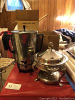 Vornado coffee maker next to metal serving dish with lid and warming candle stand on red surface, wood-paneled background.