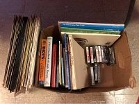 Box containing over 25 vinyl records stacked vertically alongside books and cassette tapes laid flat at top of the box