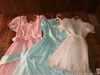 Three vintage girls' formal dresses displayed on hangers on a wooden surface: pink lace dress, blue dress, white dress.