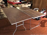 Two 6 foot folding tables shown side-by-side; one with wood grain plastic top in good condition, other with worn solid wood top, both metal-legged folding tables.