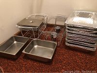 Overview of six metal Sterno food warming frames, three metal trays (one with lid), and a stack of disposable aluminum trays, arranged on carpet floor.