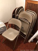 Ten metal folding chairs stacked and one chair open in front on red carpet. Nine beige chairs and one brown chair partially visible behind.