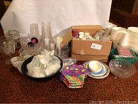 Wide view of assorted plastic cups, paper plates, napkins, trays, and bowls, some new in plastic packaging, laid out on carpet.
