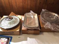 Three serving platters shown in their original boxes: Kromex Lazy Susan crudite tray with glass sections, Glassbake rectangular glass tray sealed in plastic, and silver plated cake stand wrapped in plastic.