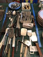 Full view of assorted cutlery and kitchen tools on table, including forks, knives, spoons, spatulas, and pizza cutter.