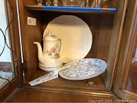 White Lenox platter with embossed floral pattern behind the coffee urn and cake plate with knife on a wooden shelf