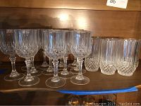 12 crystal wine goblets and 6 crystal tumblers arranged on a wooden shelf, showing the etched and faceted design of the wine glasses and the diamond cut and vertical etching on the tumblers.