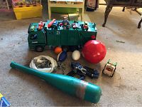 Green Bruder toy garbage truck with toy cars, balls, toy soldiers, a bat and other accessories arranged on carpet