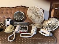 Photo showing two fans, four corded phones, one round wall clock, and one digital clock radio arranged on a couch.