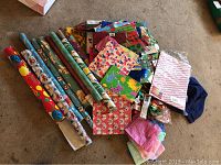Photo showing multiple rolls of wrapping paper with Christmas and holiday themes, gift bags, and tissue paper laid out on floor.