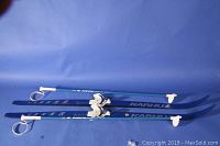 Blue Karhu cross country skis with white Pinso Nordic Norm bindings laid flat on blue background, blue ski poles with white handles.