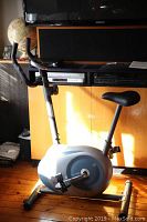 Full view of the stationary exercise bicycle showing seat, pedals, base, and flywheel housing in front of wooden cabinet.