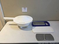 Photo of Tupperware white double colander with handle on left and two-piece Tupperware grater on right, placed on white table.