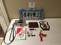 Photo of all lot contents including camera, license plate, wallet, patch, padlock, coins, keychain ornaments on a tiled floor.