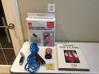 Table with home comfort weatherization kit box, blue extension cord, Black & Decker stud sensor, small utility knife blades box and 'Share The Flame' book arranged for viewing.