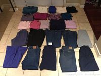 Photo showing 24 assorted ladies clothing items laid out on tiled floor. Includes 14 tops of various colors and styles along with 10 pairs of pants and jeans in various colors.