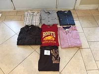Seven men's shirts and t-shirts laid out on a tile floor, including button-up shirts and graphic tees in various colors and styles.