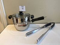 Image of Lagostina saucepan with attached glass lid and steamer insert placed on top, and KitchenAid BBQ tongs lying beside the pan.