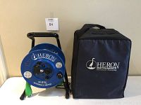 Blue circular Heron Instruments H.01L interface meter reel with attached probe, positioned next to the black soft carrying case with logo.