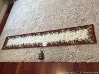 Full view of the reversed fabric holiday table runner laid out flat showing its length and floral border design with the small Christmas tree bell decoration beneath it on carpet.