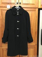 Full view of the black London Fog ladies spring/fall coat size 2X hanging on a hanger against wooden cabinets.