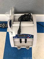 Front view of Seizer electronic money counting machine with power cord on top and remote control next to it.