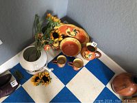 Overview of orange ceramic rooster and sunflower themed kitchenware including pitch, cups, bowls and a serving plate with artificial sunflowers and a large white ceramic dispenser.