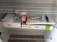 Nintendo Wii console, original box, Wii Fit Plus balance board, two Wii remotes, game cases and boxed Zumba game on wire shelving.