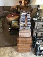 Photo showing a tall stack of various decorative baskets and storage trunks arranged in a living room setting near couches and side tables. The top items include fabric-covered boxes with faux leather trim and buckles, below is a faux crocodile textured suitcase, and the base is large woven wicker baskets.