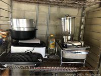 Wide view of metal shelf holding large stainless steel chafing pan, black pot, silver pot, Proctor Silex electric chafing pan, bottle of canola oil, and black electric grill.