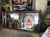 Large rectangular wood framed mirror with painted Bass Brewing Co Pale Ale logo and slogan, leaning against shelving in a storage area.