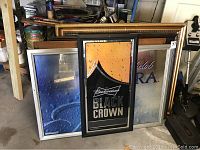 Overview photo showing multiple mirror bar signs stacked against wall and workbench, including Budweiser Black Crown and partially visible other signs