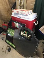 Two coolers including a large red plastic one and multiple bar mirror signs arranged in front of it. A charger and power cords are visible on the floor.
