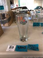 Front view of one clear Heineken 20 oz tall beer glass on table among other glassware and boxes, showing logo clearly.