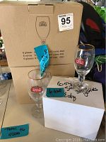 Photo shows two white cardboard boxes with Stella Artois logos indicating 6 glasses per box, two stem goblets in front, one on the box and one on a white surface with handwritten notes about the glass count.