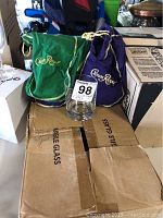 Crown Royal glass placed in front of two Crown Royal velvet bags, one green and one purple, showing bags' logos and drawstrings.
