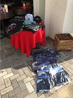 Overview of assortment of shirts on red table with some packed on ground, and two hats on top with some accessories.
