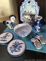 Blue and white ceramic items including figurine, vase, bowl, decorative plates, shoes, and a wall plaque displayed on blue cloth