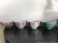 Four small Asian bowls lined up, three white with pink flowers and one turquoise with multi-color traditional designs.