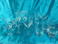 Lot of various clear glassware pieces including champagne flutes, martini glasses, highball glasses on blue fabric background.