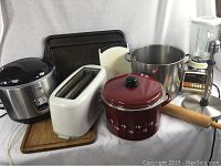 Various kitchen appliances and utensils displayed on white background including a stainless steel rice cooker, white toaster, red enameled pot, rolling pin, baking sheets, blender and immersion blender.