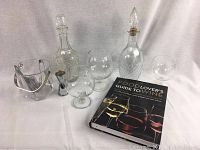Full set of barware and book arranged on white surface showing clear glass decanters, ice bucket, jigger, snifters, and wine guide book.
