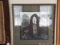 Full view of hand-painted black and white photograph framed with decorative studs along the wood frame, showing a stone arched gate with iron gate and trees behind.