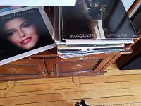 Stack of vinyl records on a wooden sideboard including identifiable album covers such as 'Imaginary Voyage' and an album featuring Lisa Manchester.