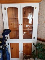 Front view of the corner cabinet showing the upper glass doors and lower natural pine wood doors with brass hinges and wooden knobs, reflecting light from the window.